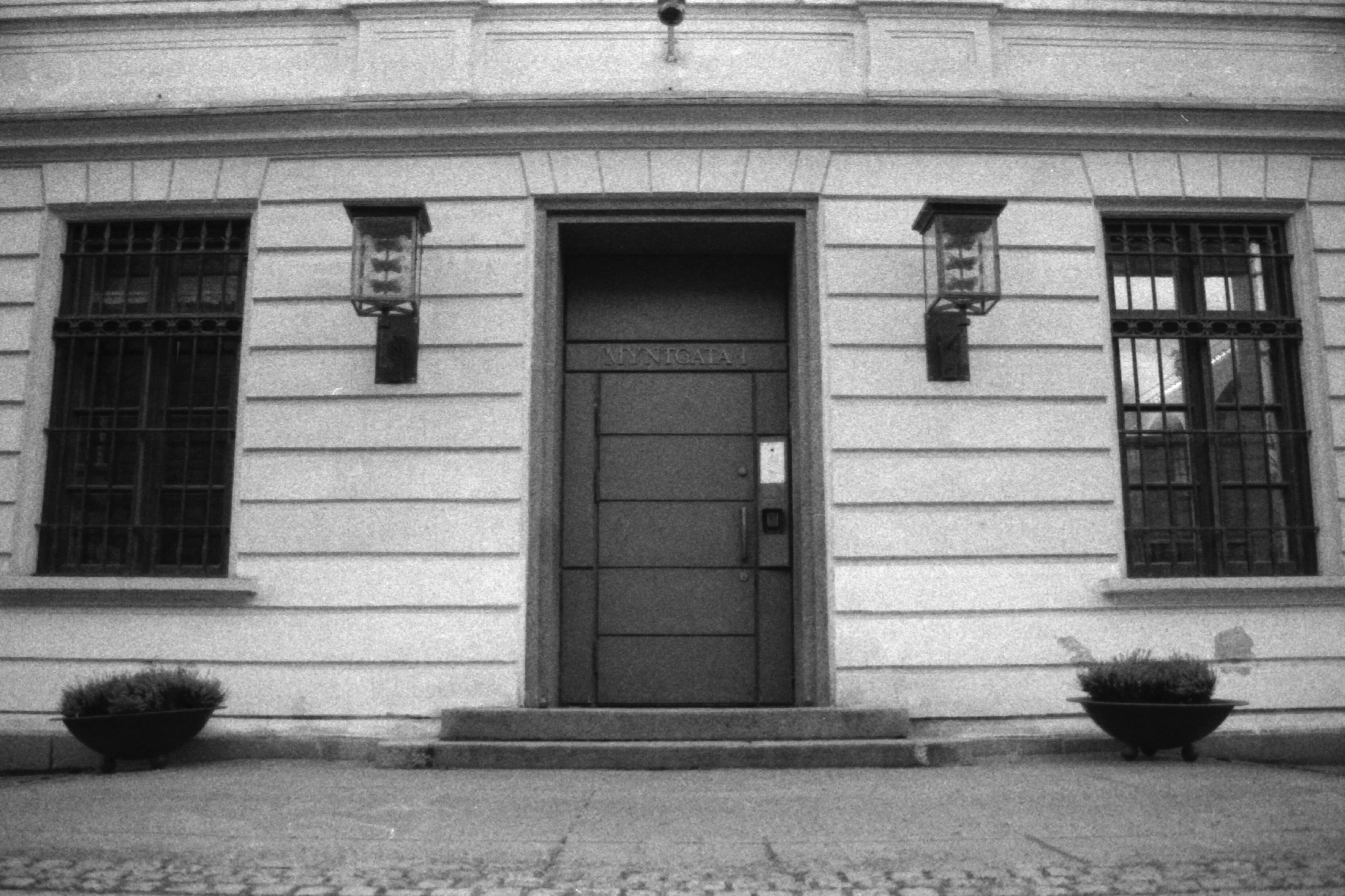Black and white picture of an entrance to a building featurintg two barred windows, two lamps and a door with the text “Myntegata 1”