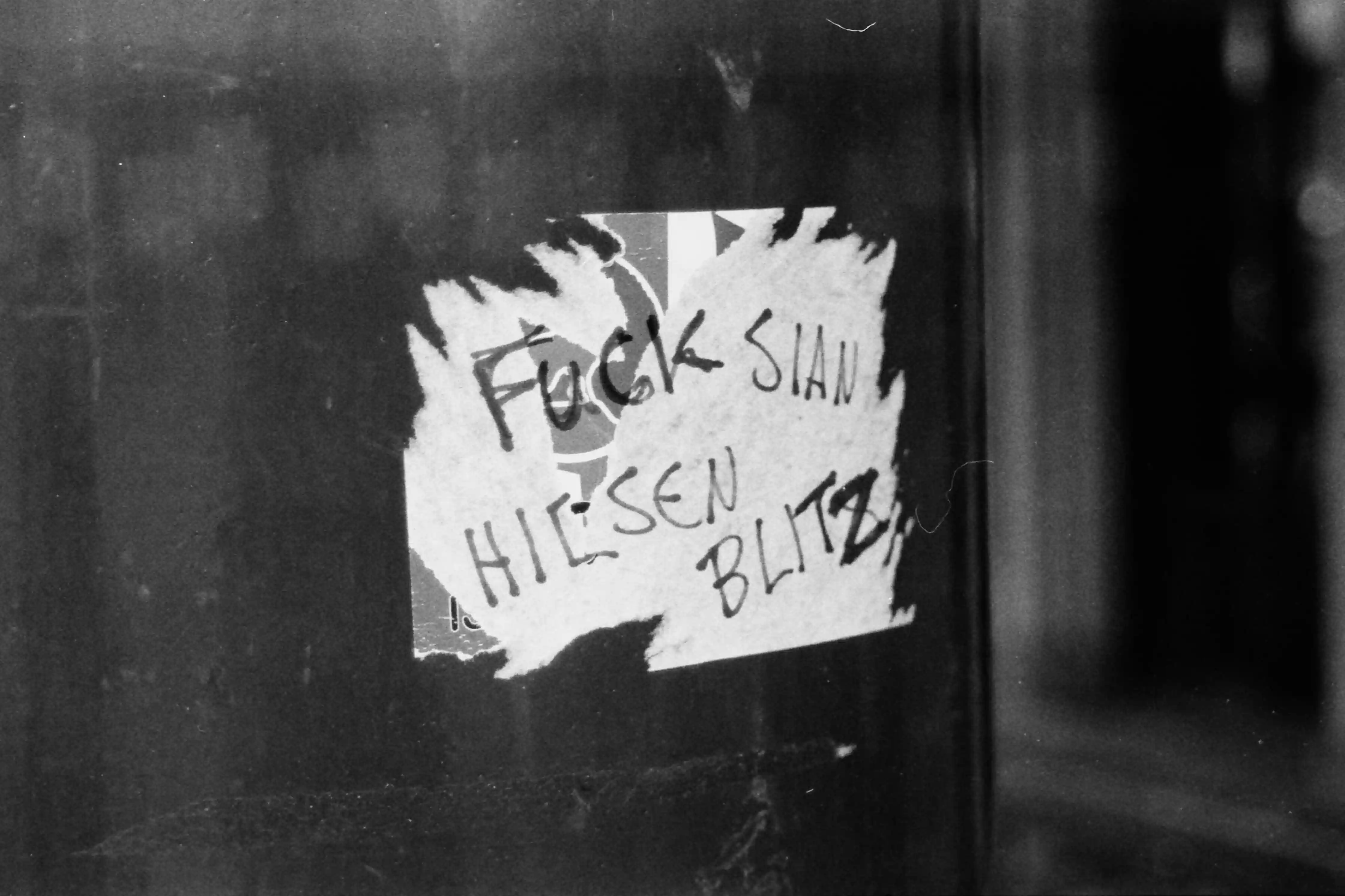 Black and white closeup picture of a partially torn sticker with the text “Fuck SIAN Hilsen Blitz”