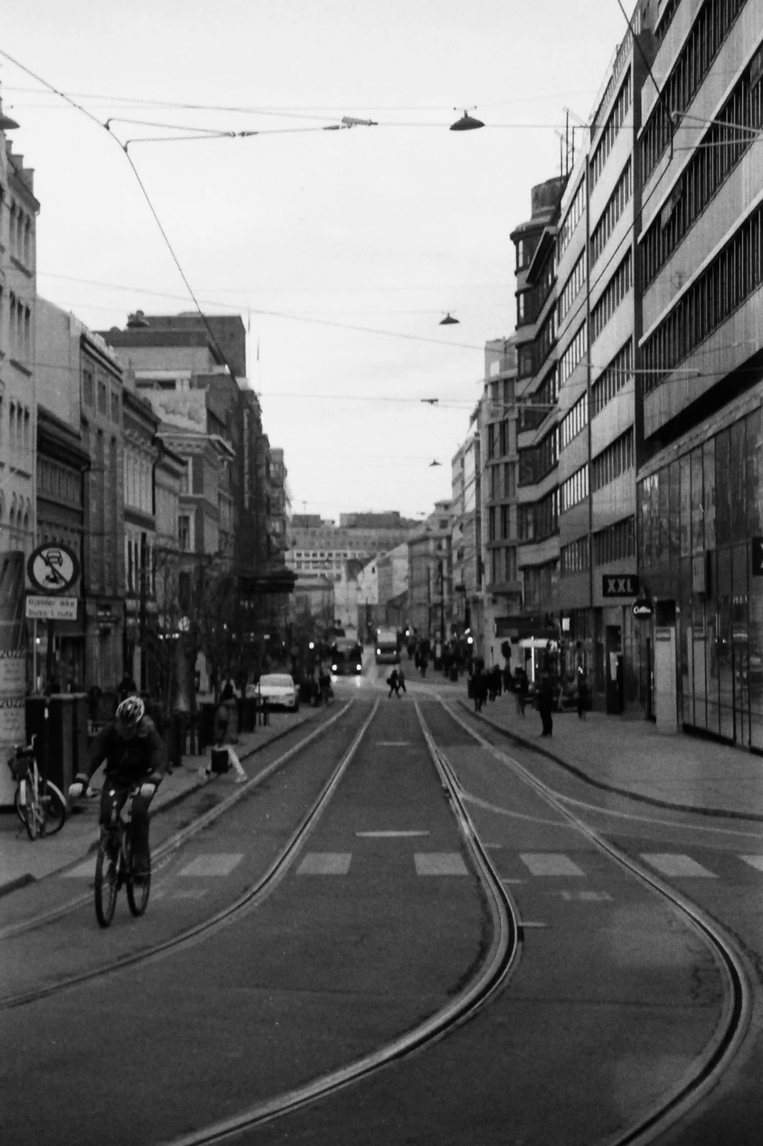 Black and white picture in portrait mode of a street featuring people on bicycles, tram tracks and some people. There are wires hanging from some poles and buildings