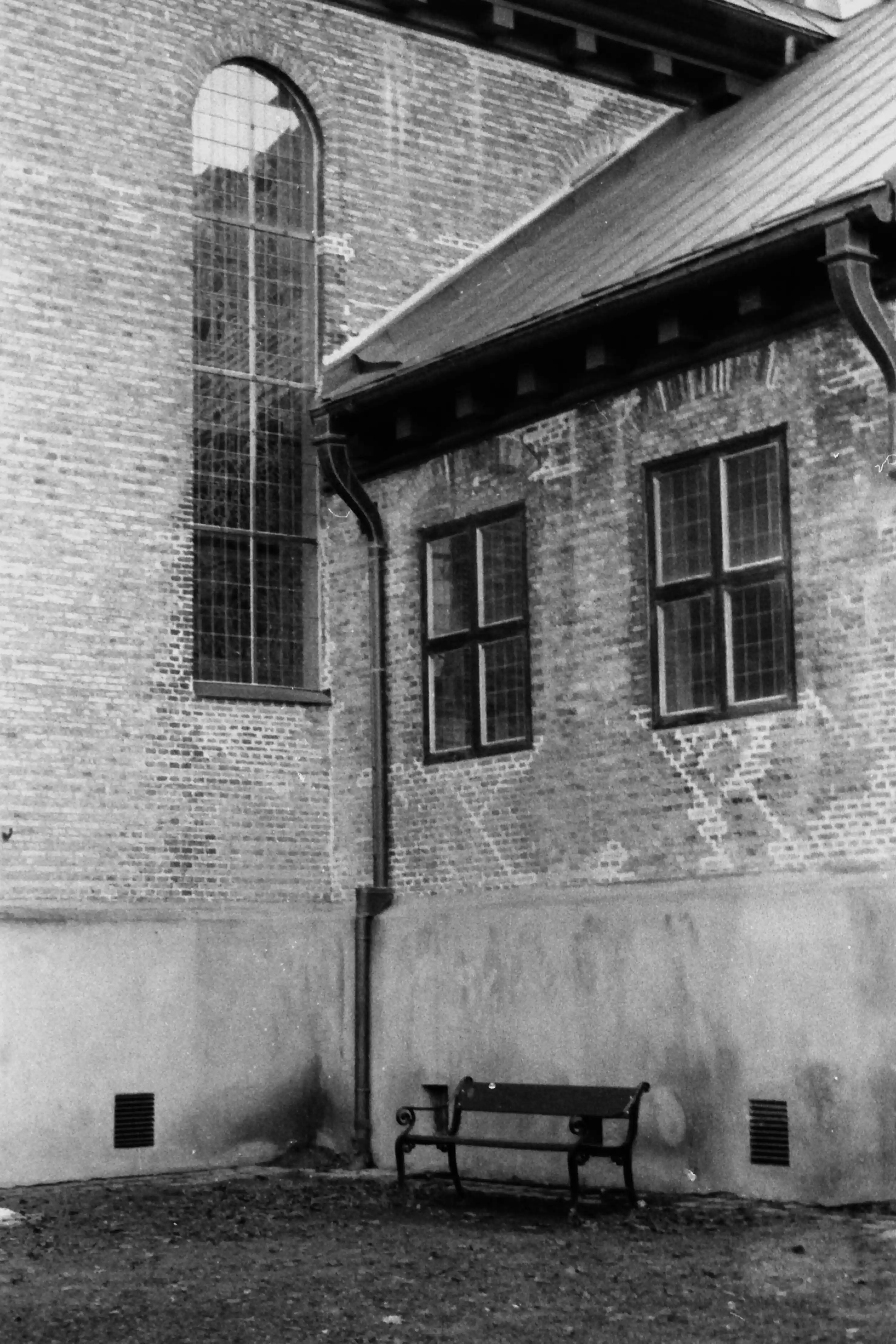 Black and white picture in portrait mode of the side of a church with a single lonely bench