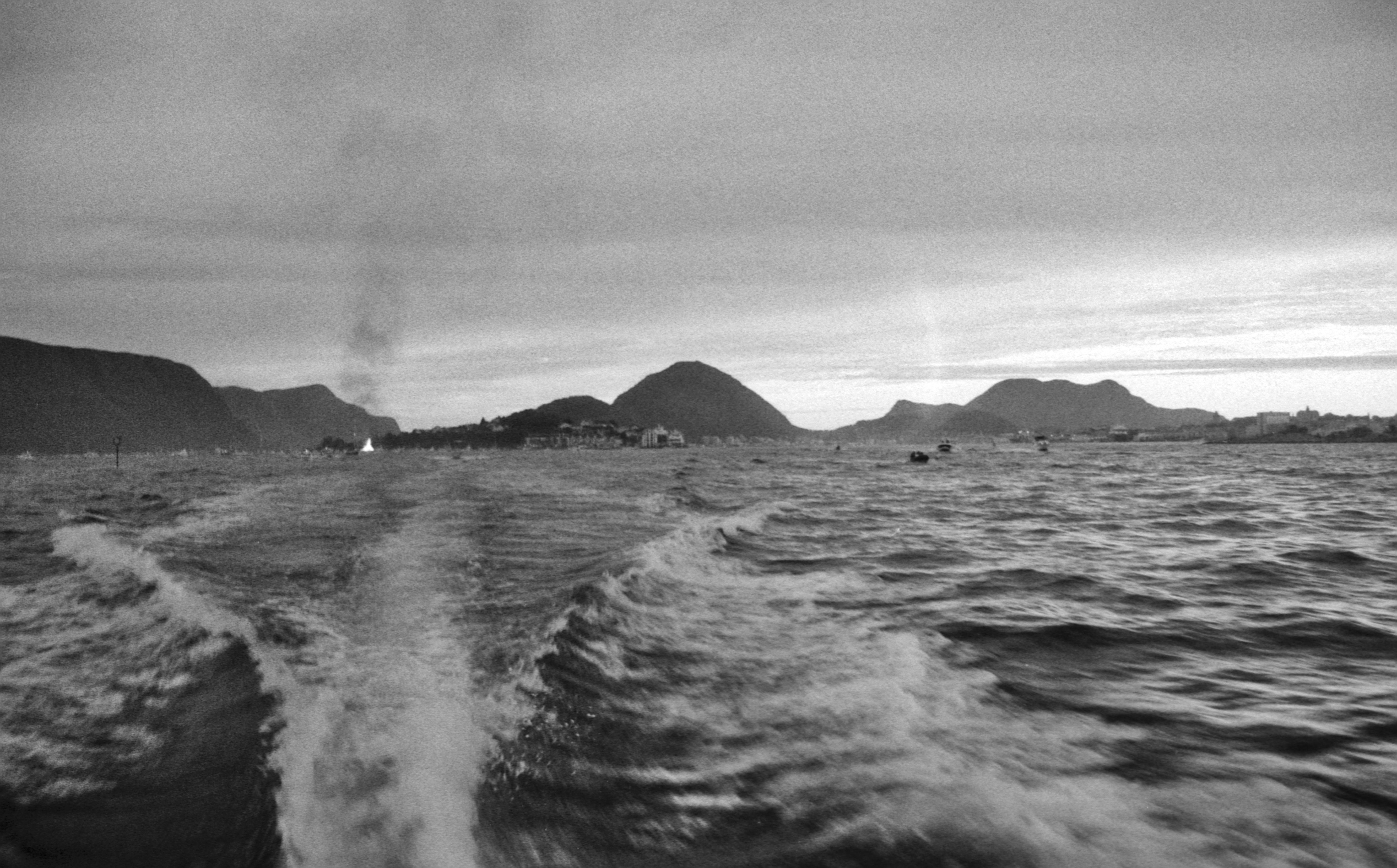 Black and white picture of the sea and some mountains along the coast taken from the back of a boat so you can see the speed waves